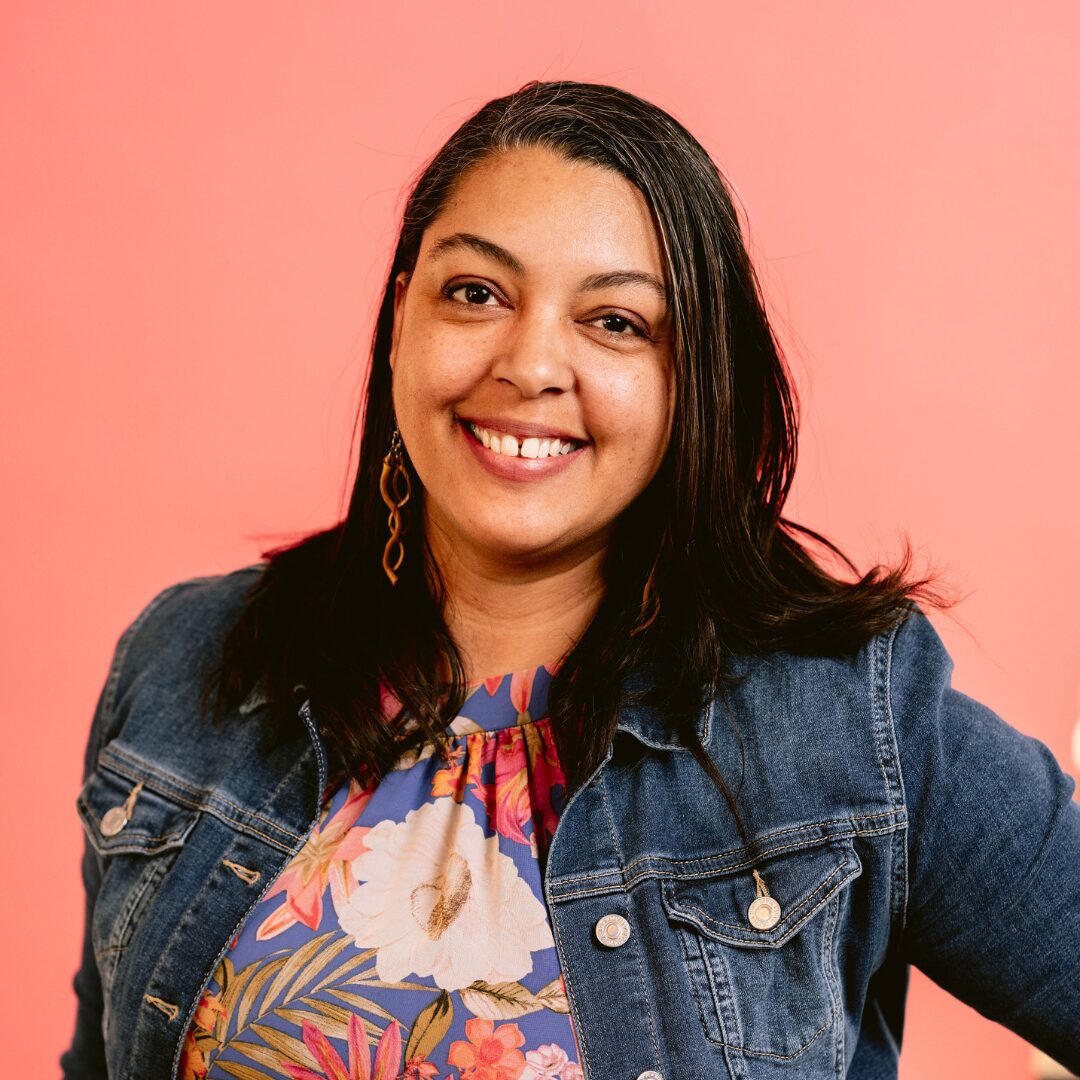 A picture of a mixed race woman with shoulder length brown hair. She is wearing a floral top and a jean jacket and is in front of a salmon colored backdrop.