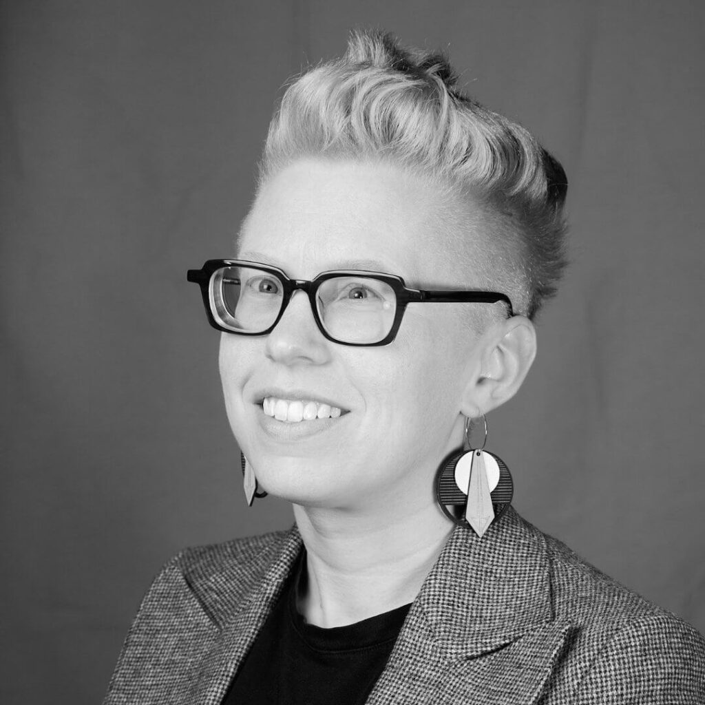 Megan Eckman, black and white photo, smiling while wearing glasses. She has short hair, styled super cool, and great earrings!